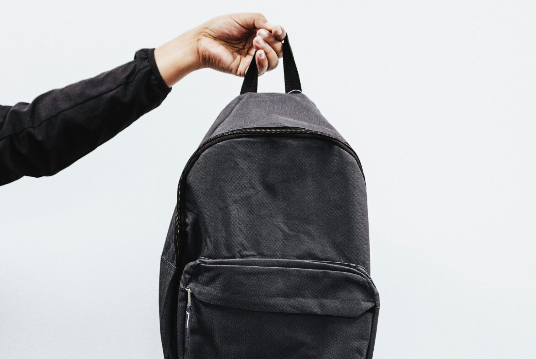 A simple black backpack being held by a hand against a minimal white background, emphasizing style and practicality.